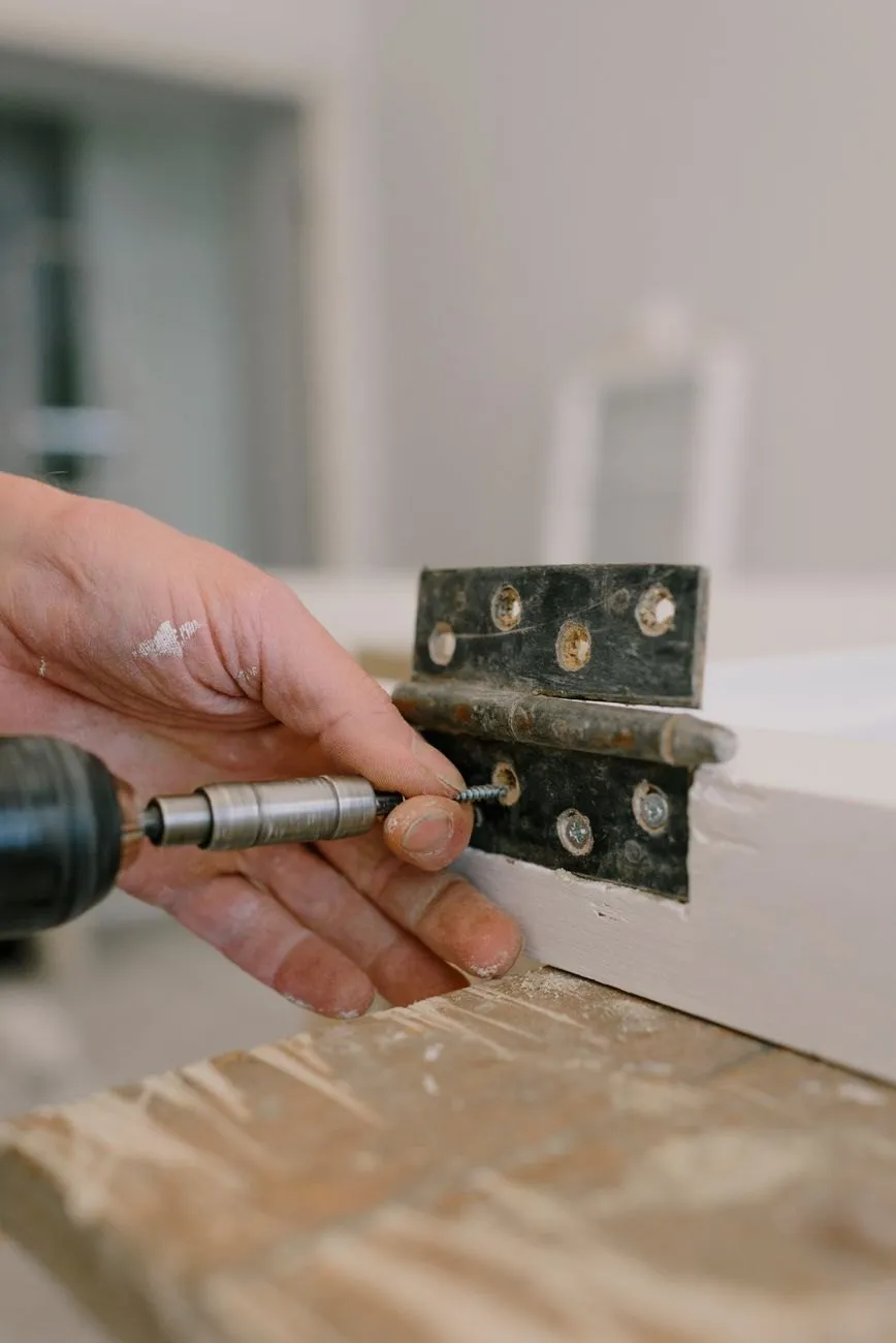 Hands holding a steel hinge against a door edge while a drill drives the screw